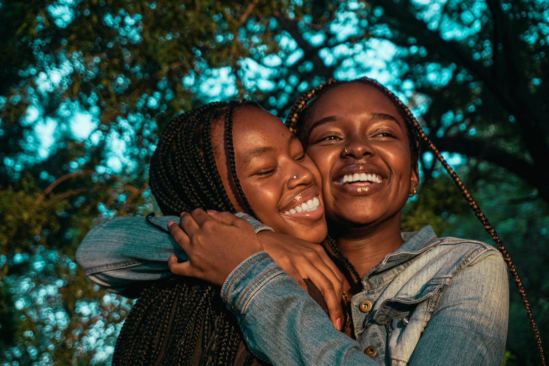 Two women in a close embrace under dappled tree light, smiling.